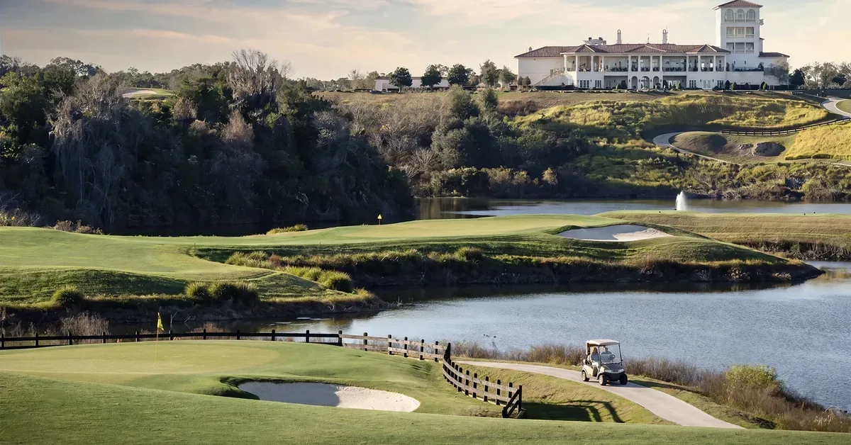 Luxury golf course estate perched on a hilltop overlooks manicured fairways, sand bunkers, and a serene lake with a golf cart on the winding path below.