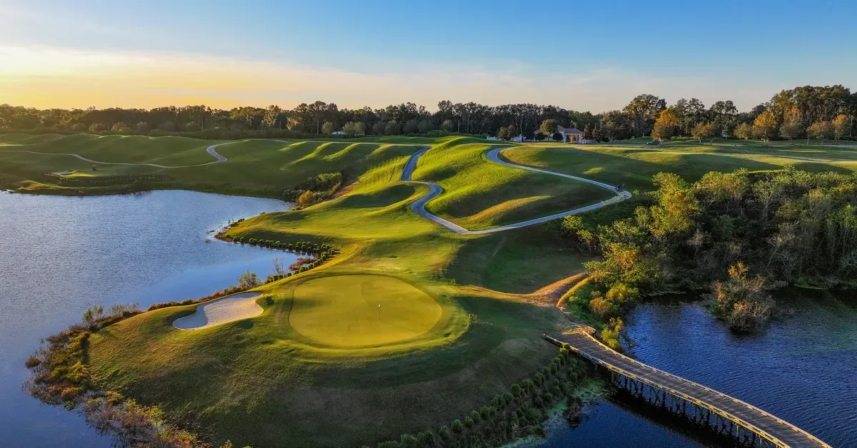 Aerial sunset view of a lush golf course with rolling fairways, sand bunkers, and a wooden bridge spanning a serene lake surrounded by trees.