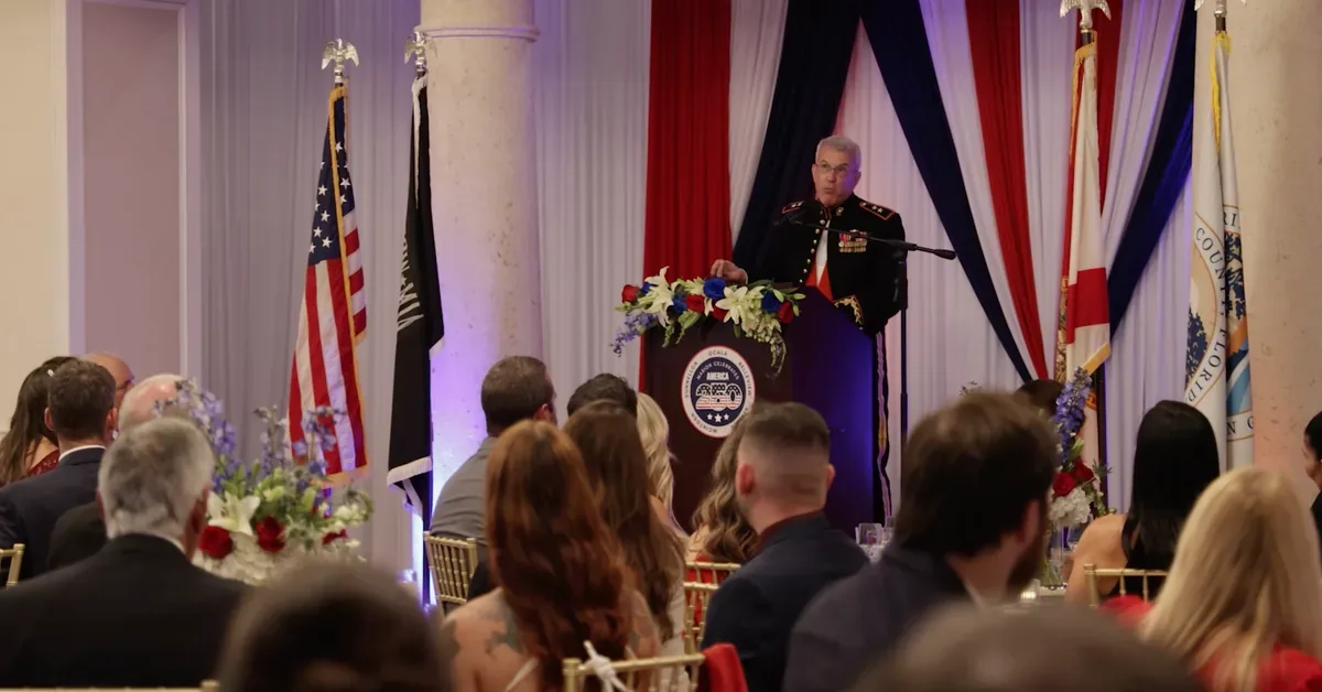 A uniformed military officer delivers remarks at a formal patriotic banquet, flanked by American flags and red, white, and blue décor.