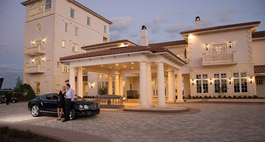 Illuminated clubhouse exterior at The Club at Iron Lake at dusk, with a columned entrance and two people beside a black luxury car.