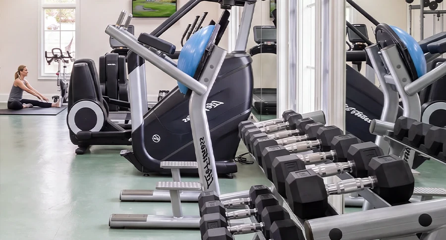 Fitness center at The Club at Iron Lake with cardio equipment, free weights, and a member stretching near the windows.