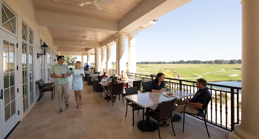 People dining and talking at tables on a covered clubhouse terrace with stone columns, overlooking a green golf course and lake.