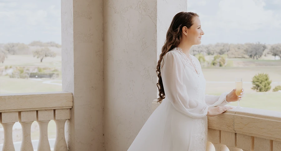 Bride in a white robe holds a drink on a clubhouse balcony overlooking the grounds in Ocala, Florida.