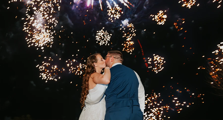 Newlywed couple kisses beneath a colorful fireworks display during an evening wedding celebration.