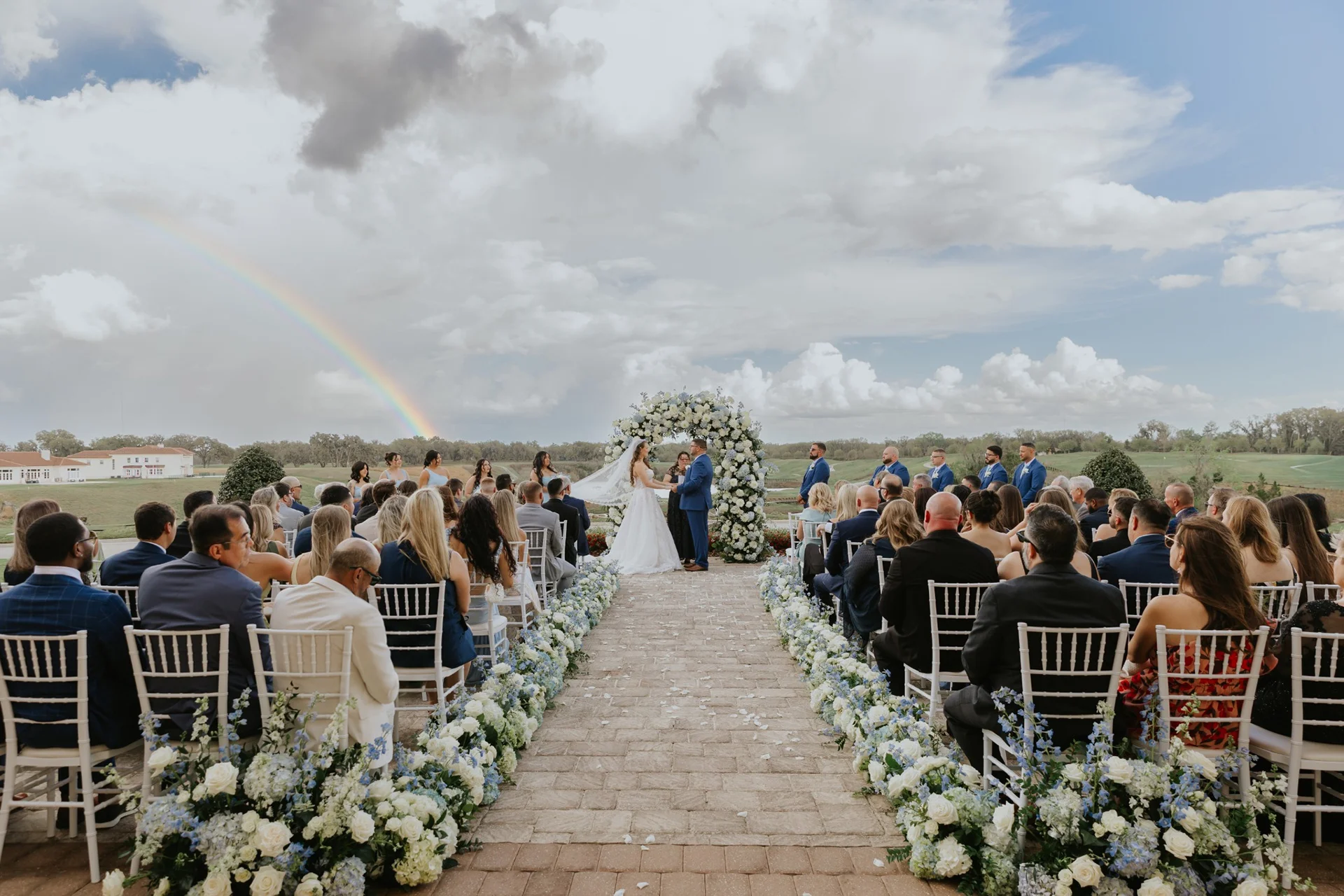 Bride and groom holding hands beneath a white floral arch at an outdoor ceremony, seated guests lining a flower-edged brick aisle, with a rainbow in the cloudy sky over a golf course.